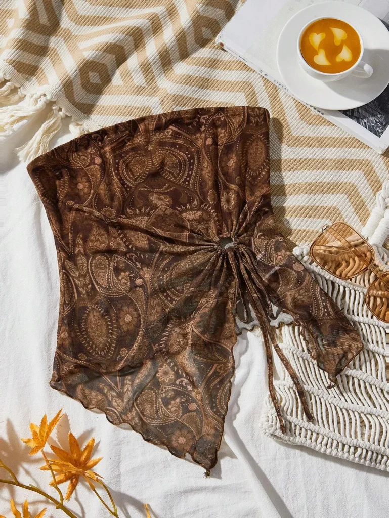 เสื้อครอปสายเดี่ยวเกาะอกสีน้ำตาลลาย paisley ที่วางอยู่บนผ้าปูที่นอนสีขาว มีถ้วยกาแฟวางอยู่ข้างๆ พร้อมแว่นกันแดดและกระเป๋าถือทรงตาข่ายสีขาว