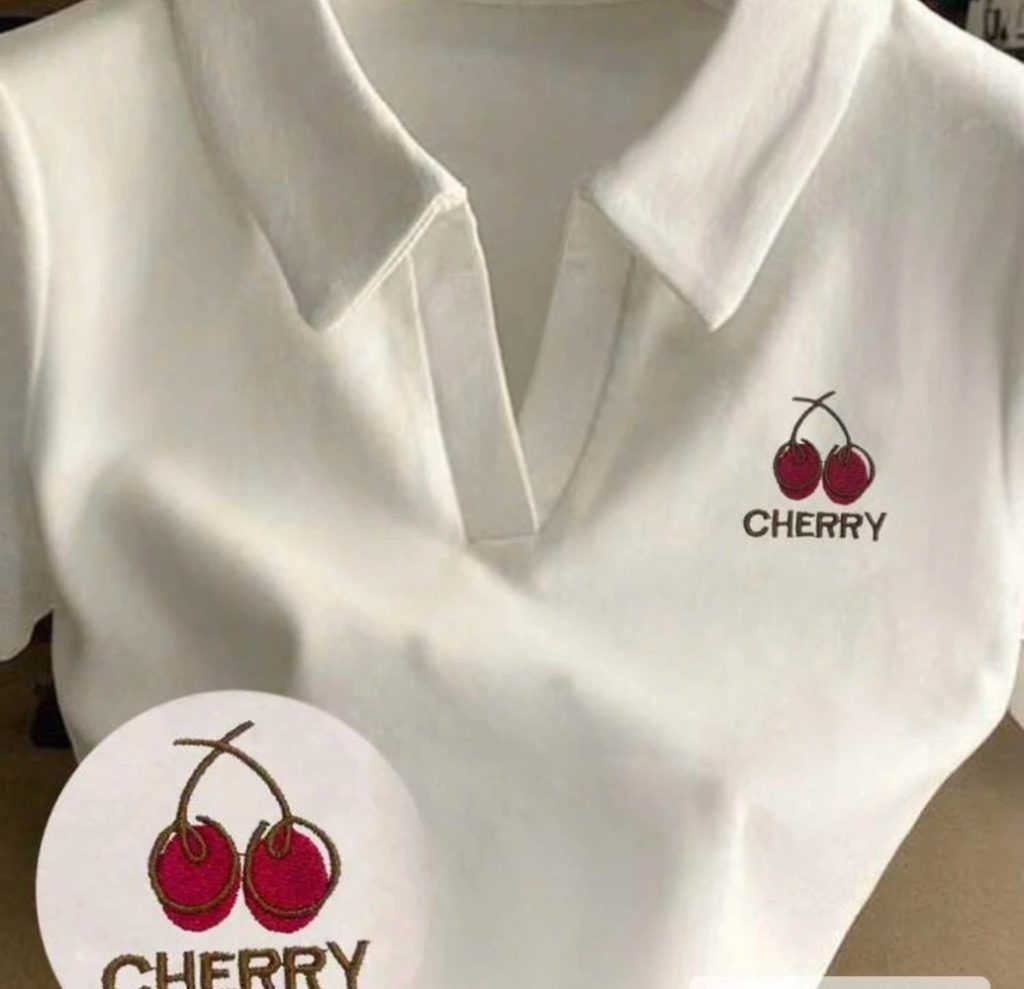 เสื้อโปโลสีขาวแขนสั้น ที่มีปกแบบคอวีลึก ปักลาย เชอร์รี่สีแดงเข้ม พร้อมคำว่า "CHERRY" สีน้ำตาลเข้มอยู่ด้านล่างลายปัก ลายปักอยู่ทางด้านขวาบนของตัวเสื้อ มีวงกลมเล็กๆ ที่มุมล่างซ้ายแสดงภาพขยายของลายปักเชอร์รี่

