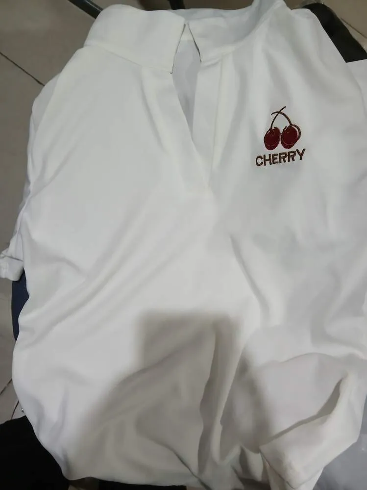 สื้อเชิ้ตหรือเสื้อเบลาส์สีขาวเรียบง่ายวางอยู่บนพื้นผิว มีปกและคอเสื้อรูปตัววี (V-neck) บริเวณหน้าอกด้านซ้ายบนมีงานปักขนาดเล็กเป็นรูป ลูกเชอร์รี่สองลูกพร้อมก้าน และคำว่า "CHERRY" ที่ปักด้วยด้ายสีแดงเข้มหรือสีม่วงแดงอยู่ด้านล่าง เนื้อผ้าดูนุ่มและค่อนข้างพลิ้วไหว

